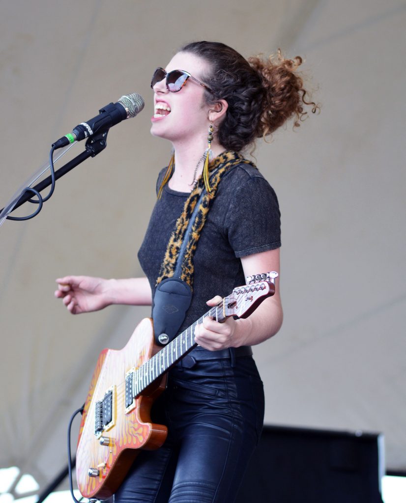 a white woman with hair in a large wild bun sings into a microphone while playing guitar.