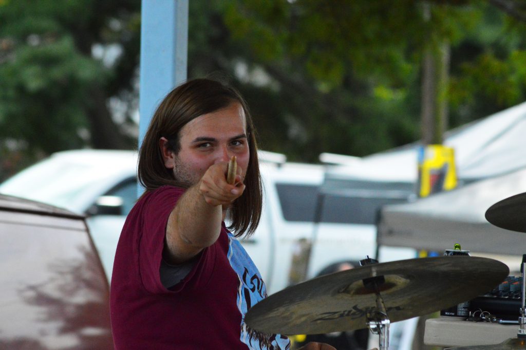 A long haired man points a drum stick at the camera. He is seated in front of a drum kit on stage.